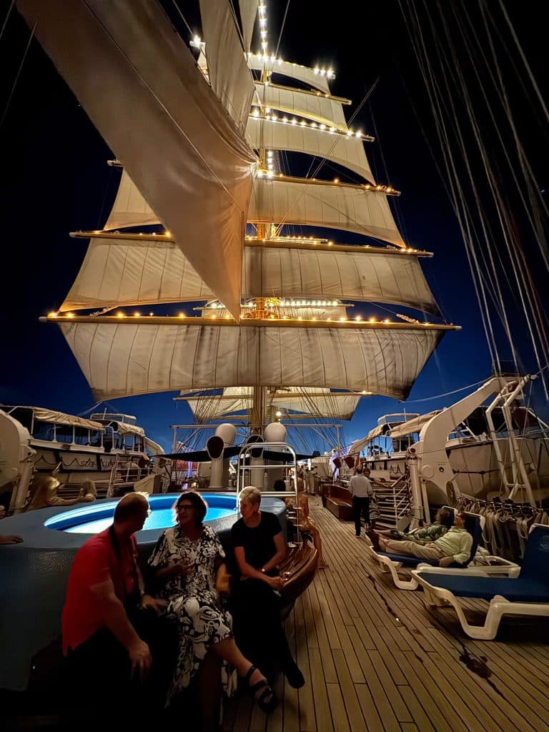 The top deck of the ship, the sails illuminated with bright lights, and a few older women sitting down on a bench.