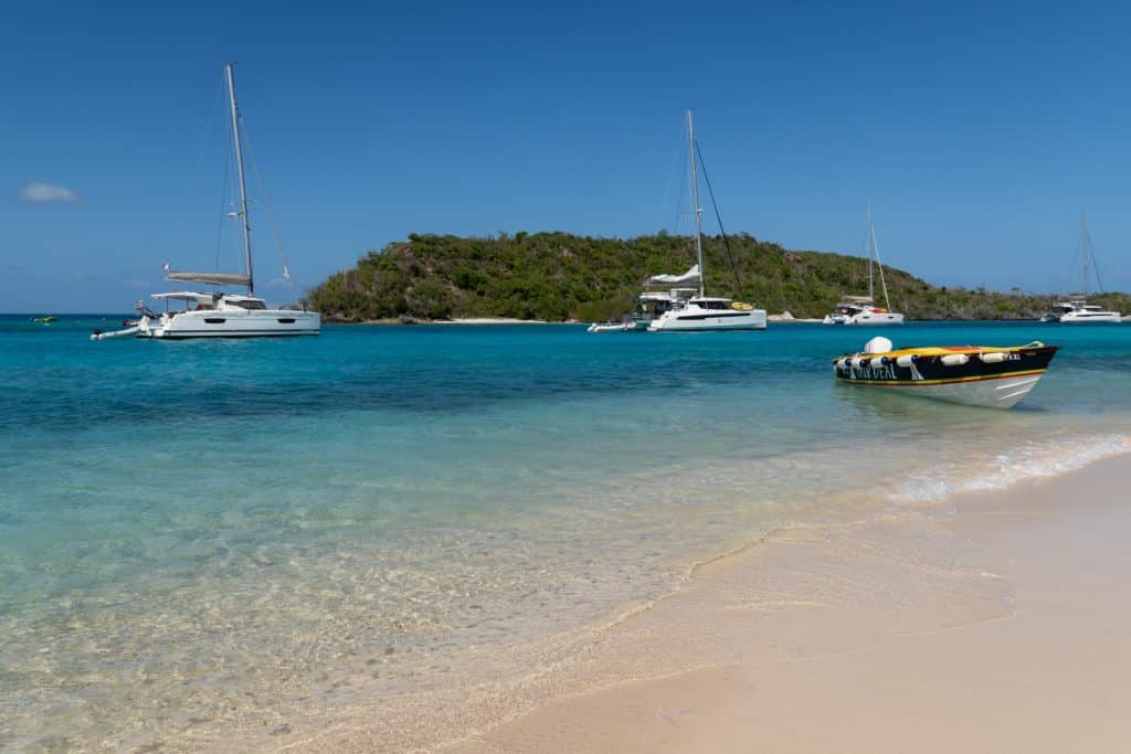 A pristine, calm beach with bright turquoise water and a few sailboats at anchor.