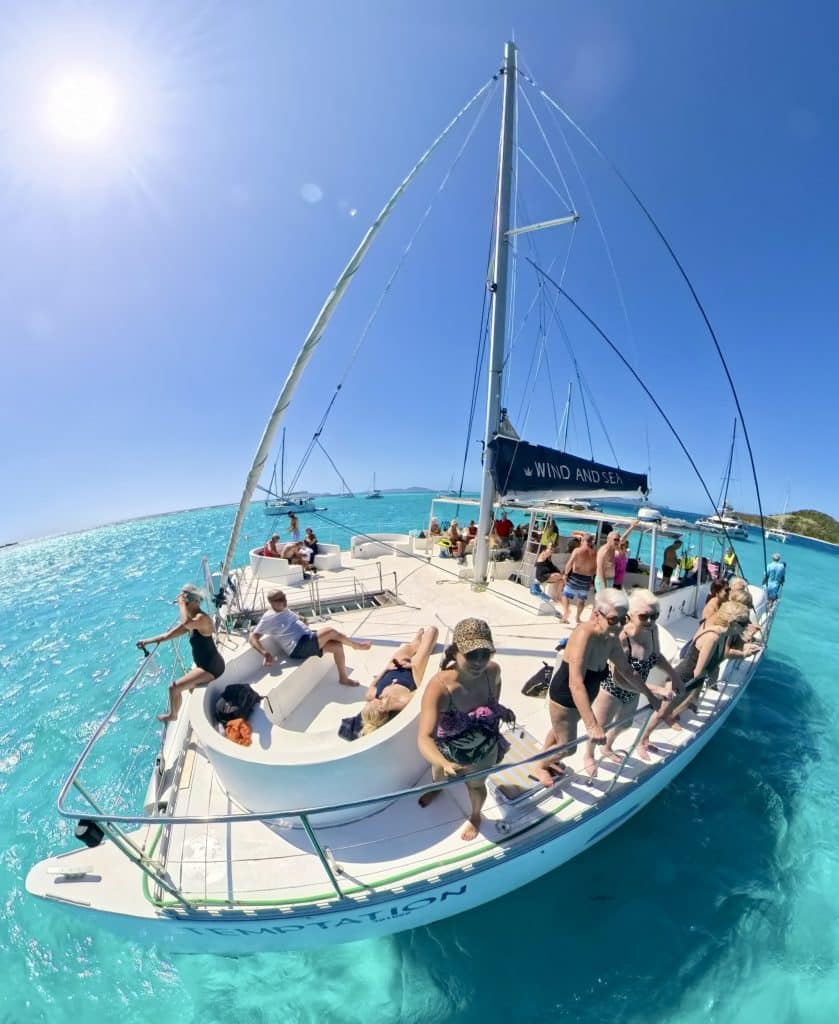 A fisheye-lens shot of Kate standing on the bow of a catamaran with lots of people behind her, sailing over the brightest turquoise water.