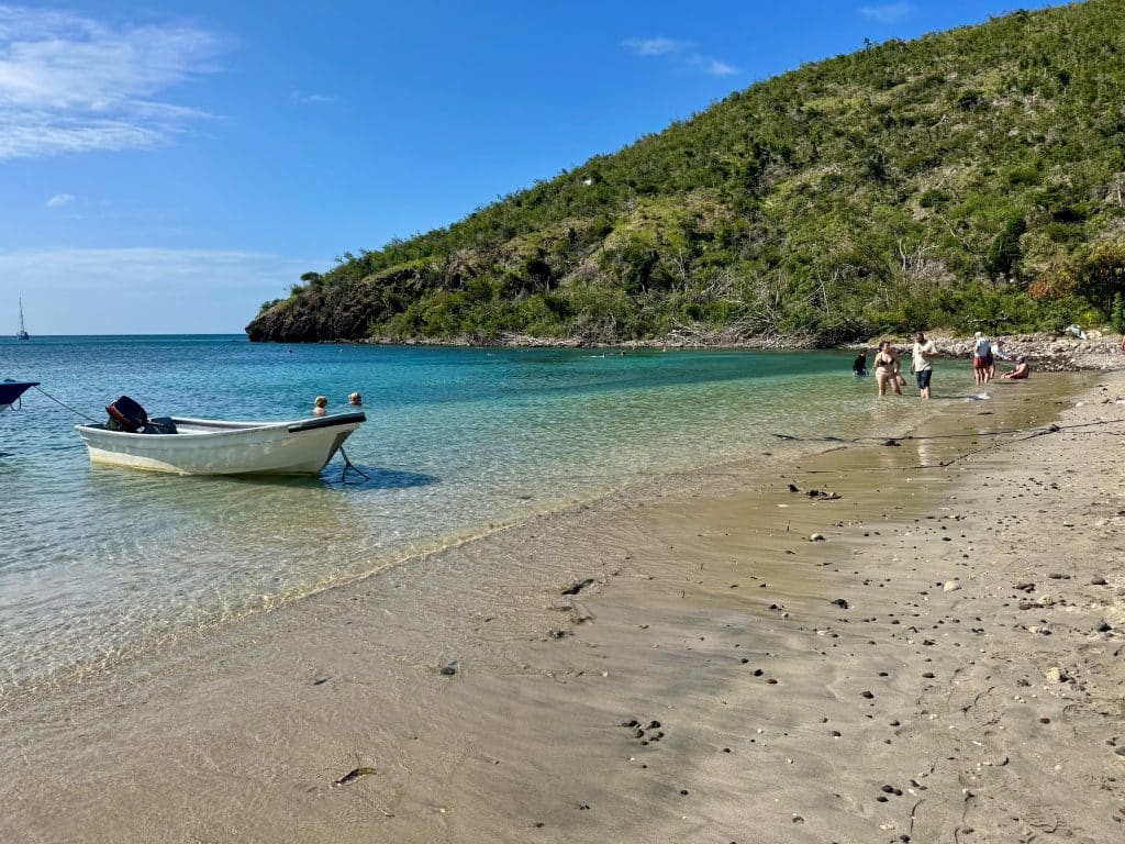 A small sandy beach with a lot of stones, and people walking down the shore to a snorkel spot in the distance.