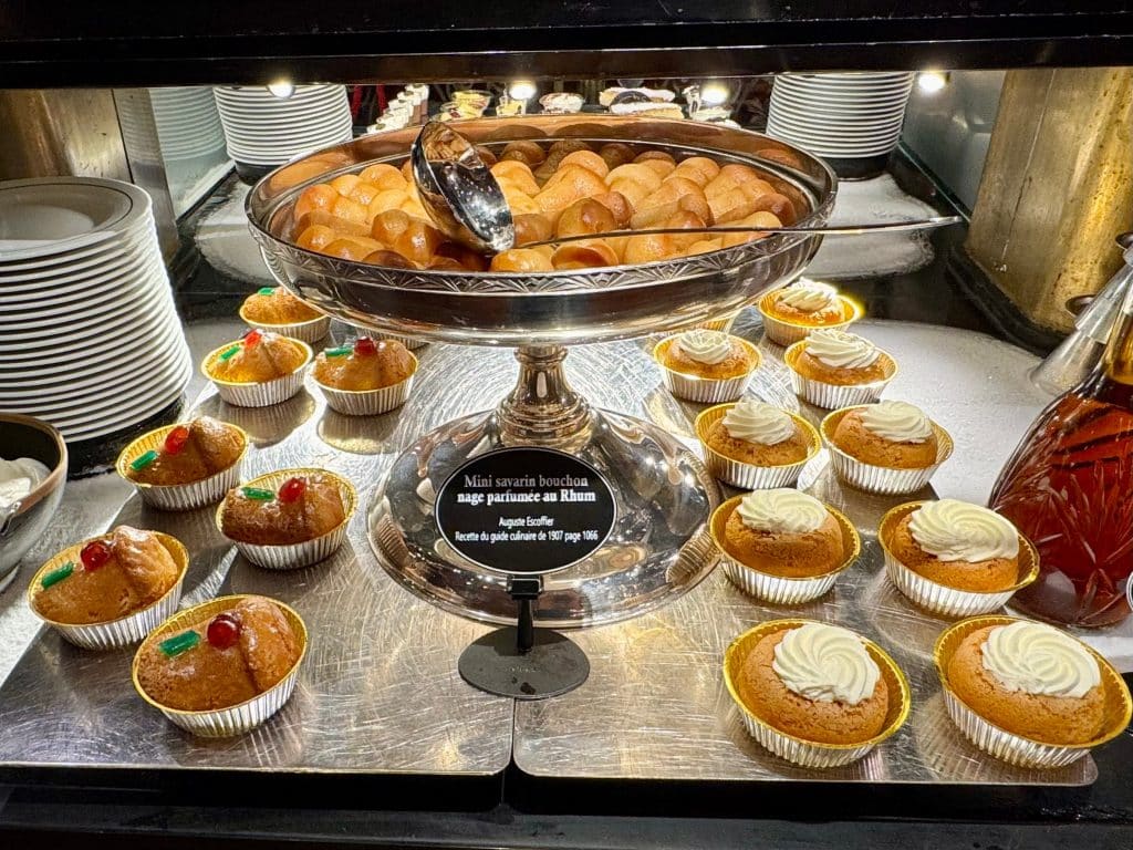 A collection of mini pastries on platters, including a giant silver bowl full of baba au rhum pastries.