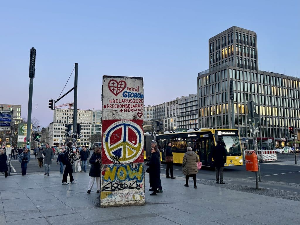 A Berlin street corner at twilight, with a big chunk of the Berlin Wall covered with peace signs and graffiti, and glittering office buildings in the background.
