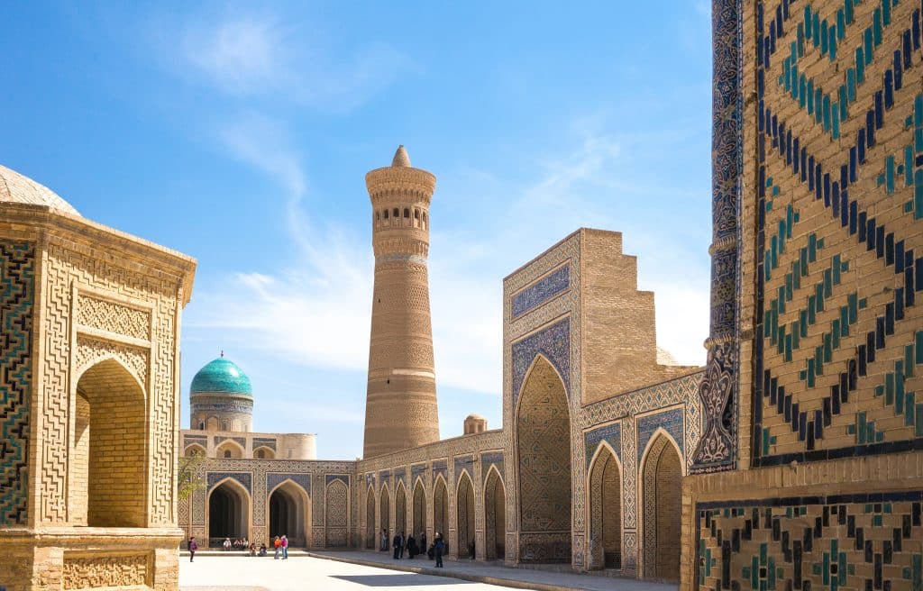 A calm square in Uzbekistan, with several towers and arched buildings the color of sand, with turquoise etching and domes.