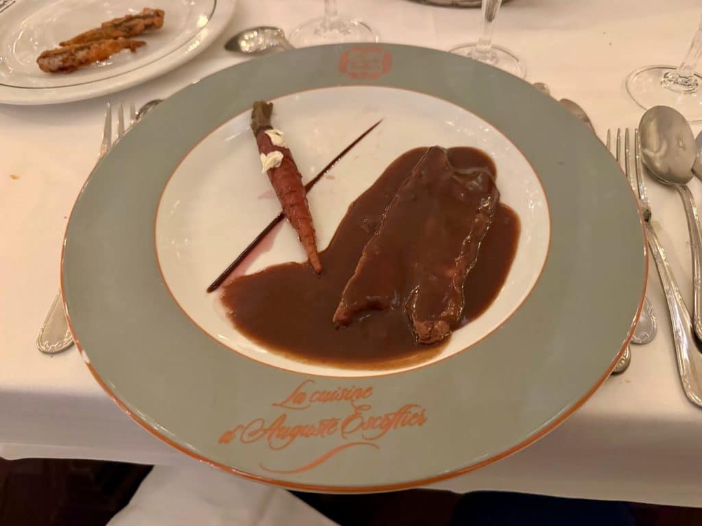 A plate with a slice of duck in a dark brown sauce, and a single skinny carrot.
