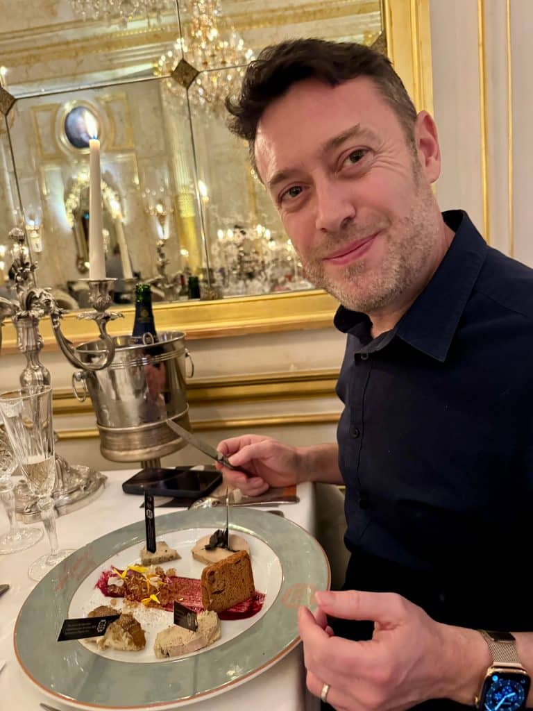Charlie smiles and holds a plate with five kinds of foie gras, each with a little tag sticking out of them.