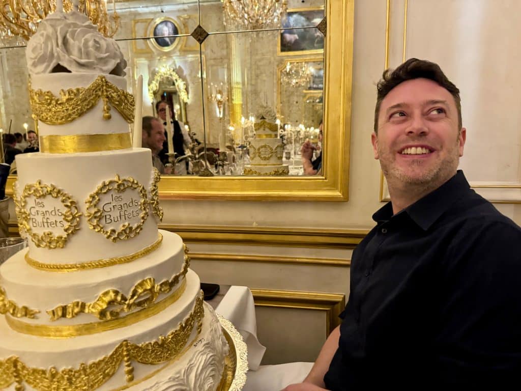 Charlie smiling wildly as a giant artificial white and gold multi-tiered cake is presented to him.