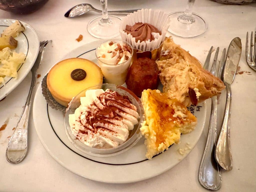 A plate absolutely covered with desserts, including a lemon tart, a chocolate mousse, a canele, a tiramisu, and a PAris-Brest cream puff cake.
