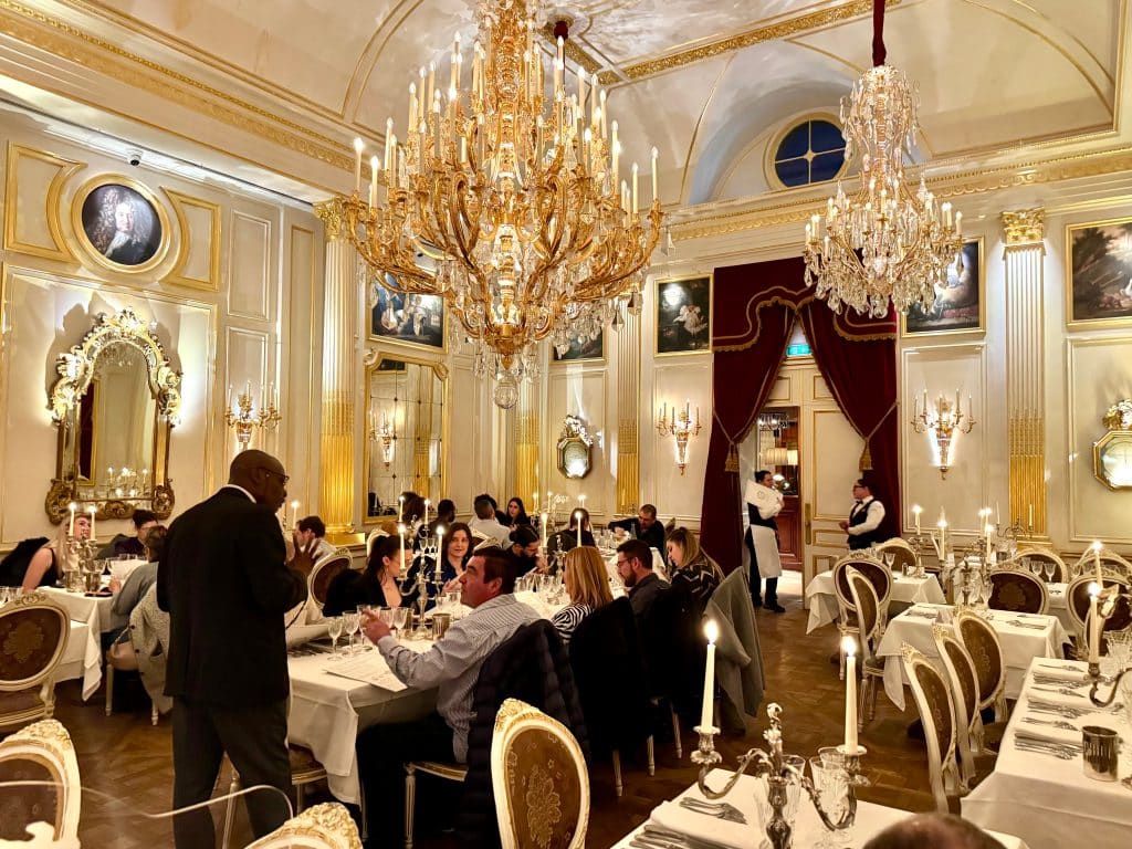 An ornate dining room with white walls trimmed with gold, and giant golden crystal chandeliers hanging overhead.