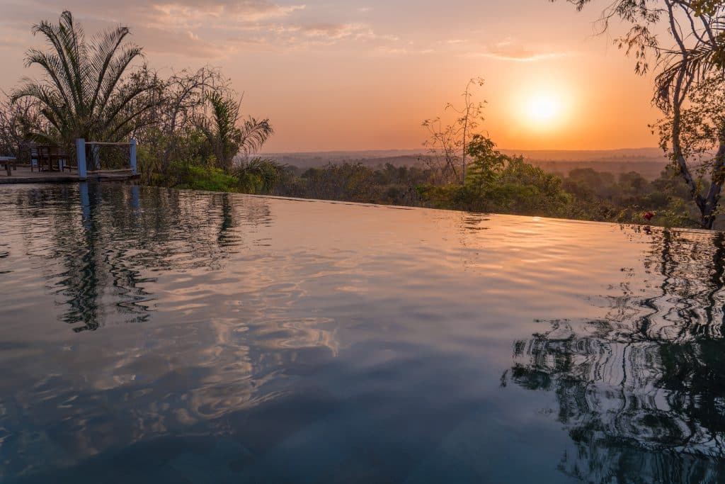 An infinity pool overlooking the jungle in the distance, with a sun setting in a pink sky.