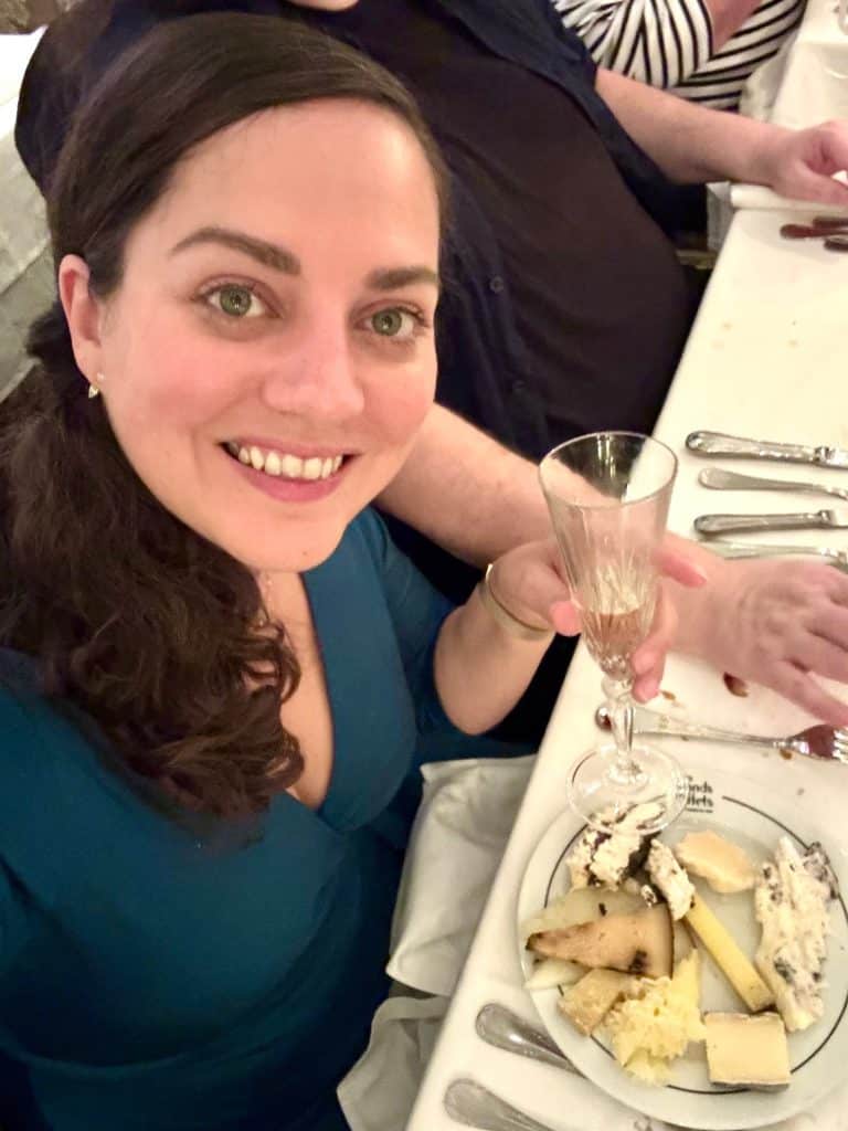 Kate taking a smiling selfie with a crystal flute of champagne and a small plate covered with around 10 different kinds of cheeses.