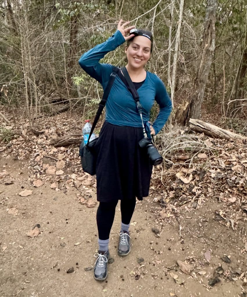 Kate wears a head lamp, camera, and wears a long sleeved shirt, dress, and leggings over sneakers while standing in the forest.