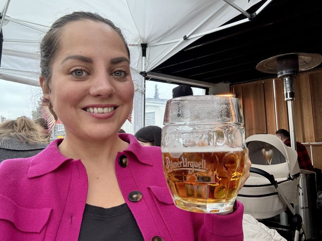 Kate wearing a pink jacket and holding up a nonalcoholic beer in a traditional Czech pilsner glass.