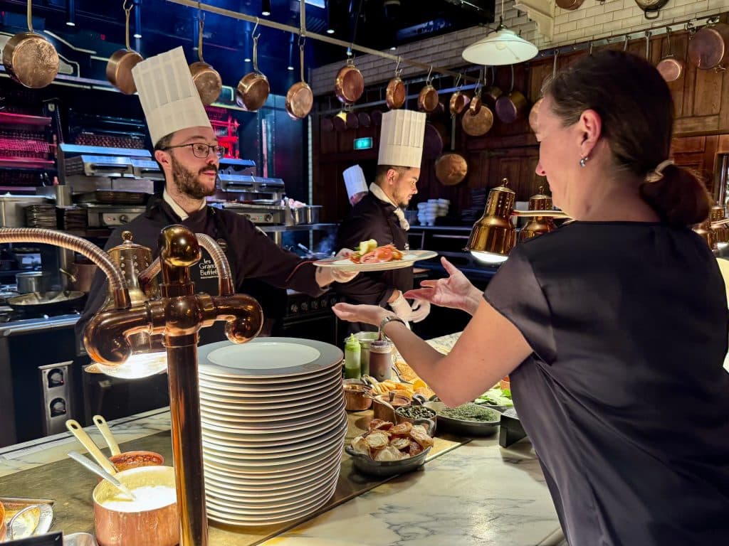 A chef in a white hat behind a counter hands Klara a plate topped with a dish.