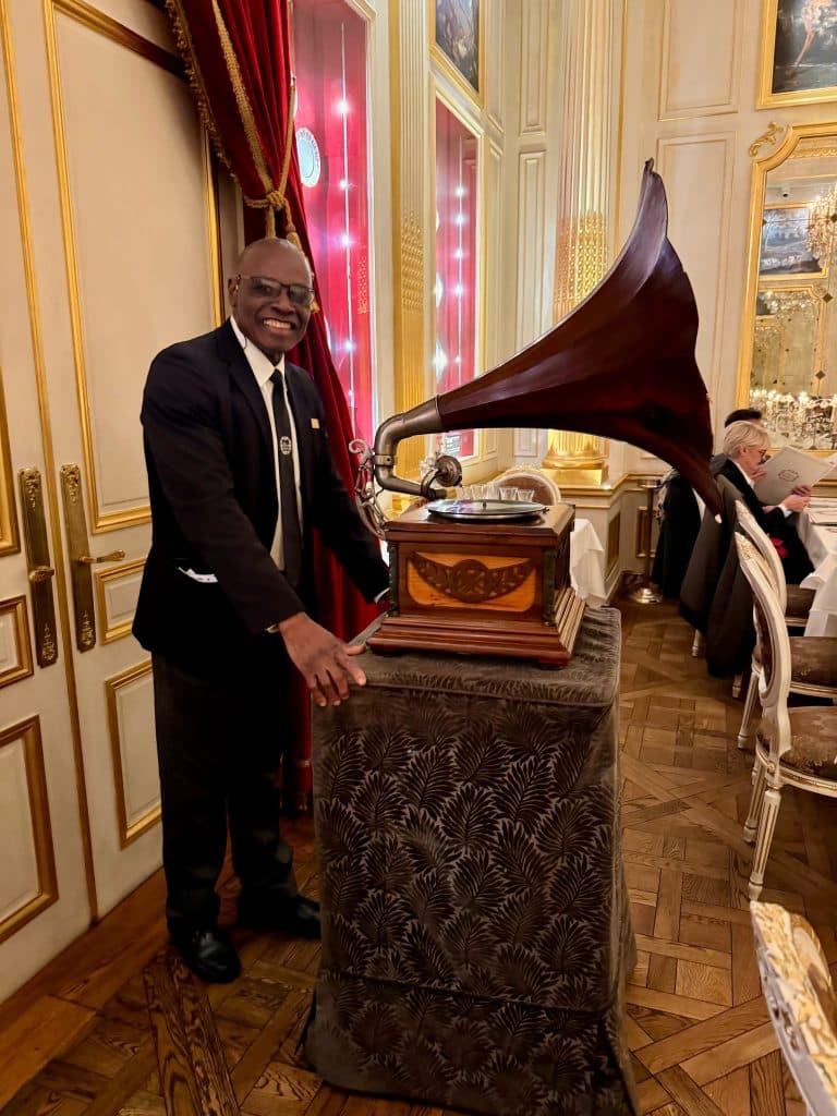 Server Leon, wearing a tuxedo, pushes a giant vintage gramophone with a huge cornucopia-shaped horn.