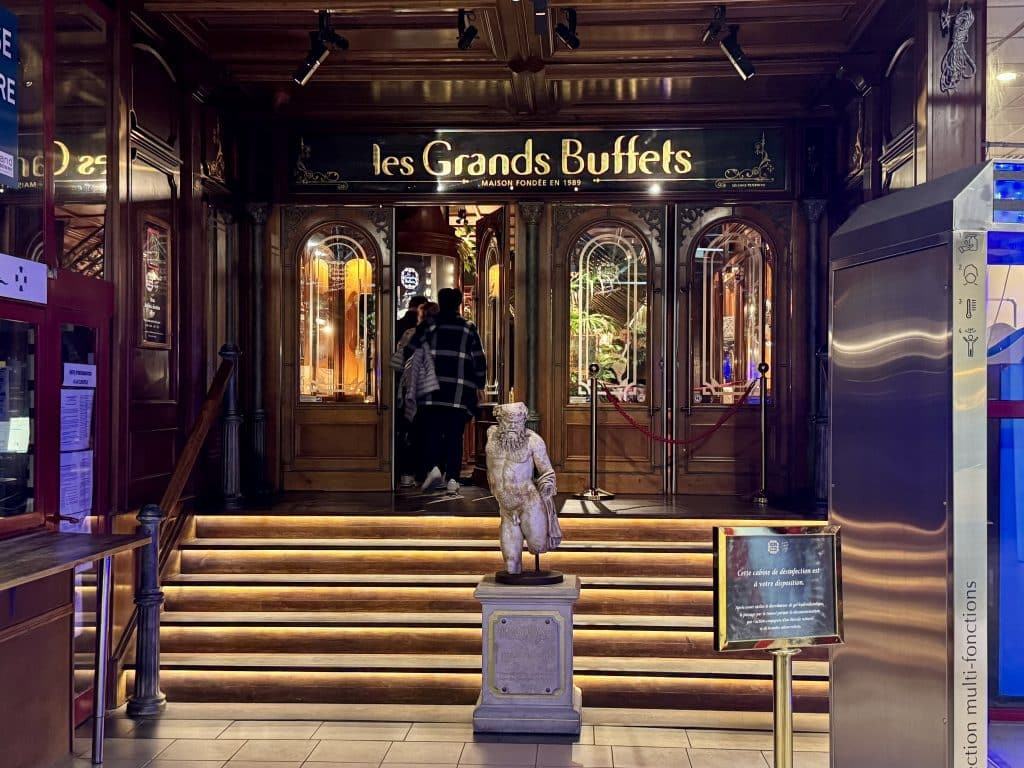 The entrance to Les Grands Buffets, with a statue in front of a small staircase leading to heavy wooden doors with glass panels in them.