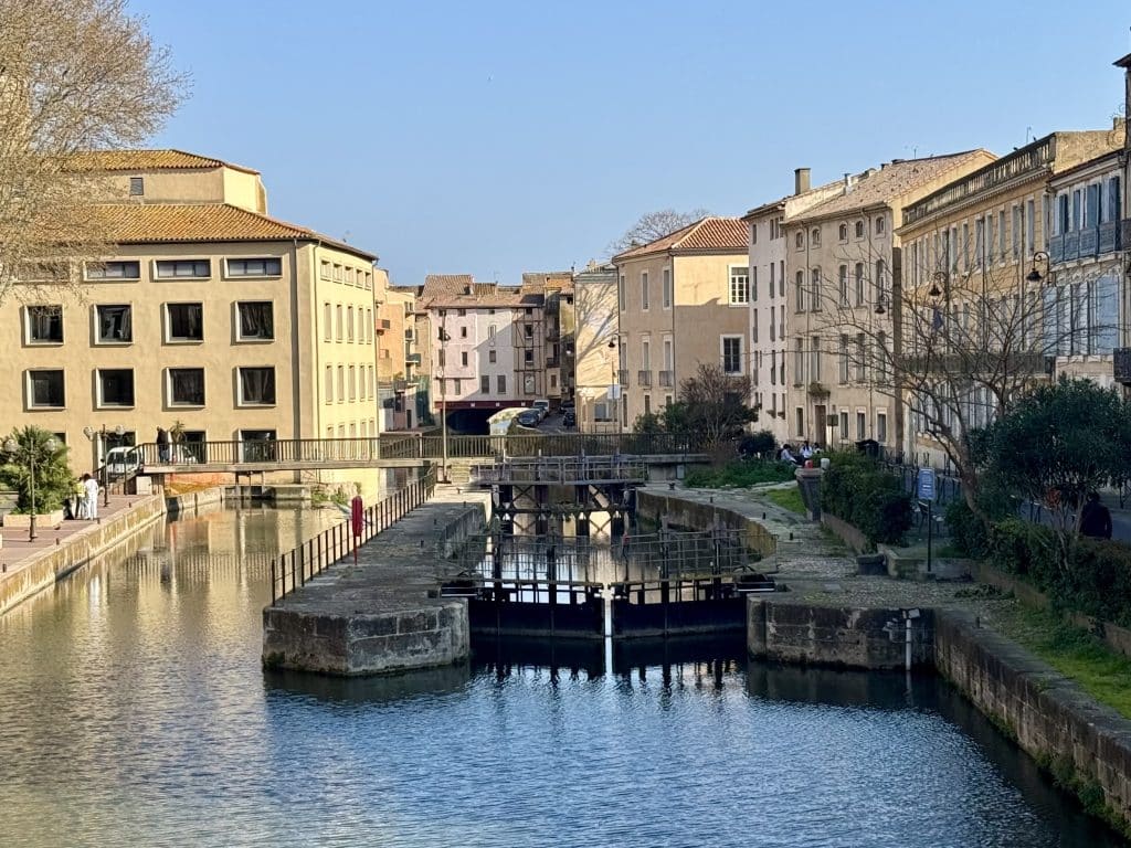 A small river canal running through the town of Narbonne, France, with pale orange buildings on each side of the canal.