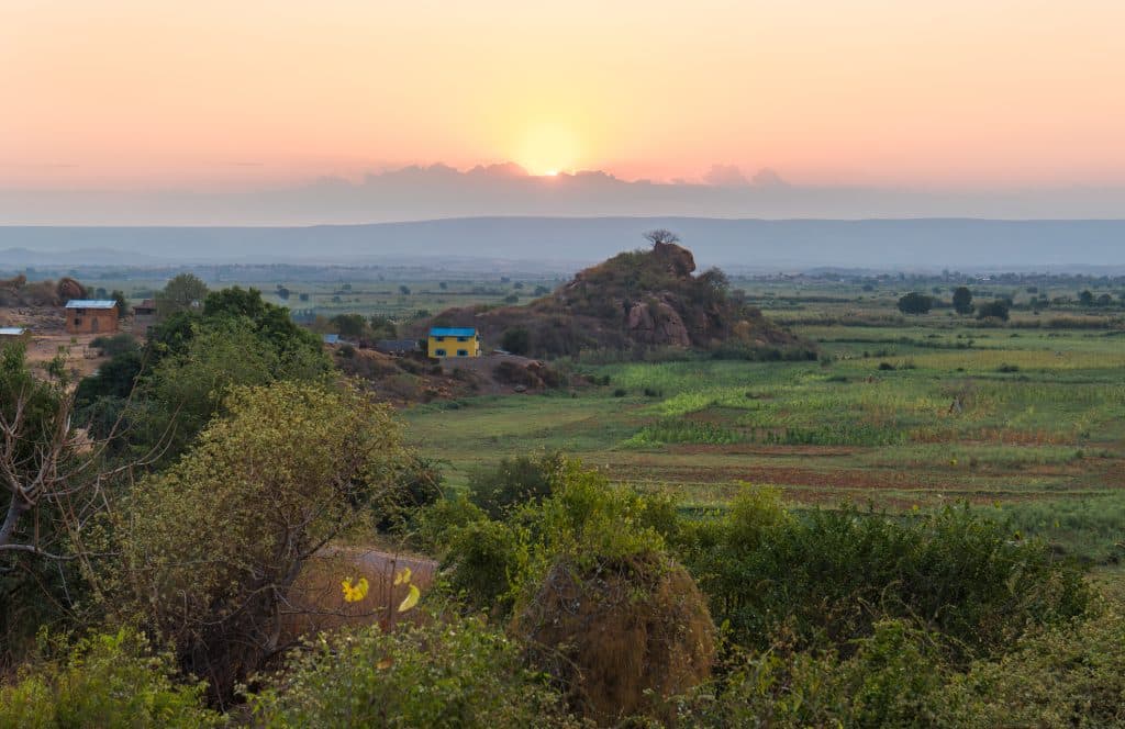 A view of endless green fields and a little yellow house with a blue roof, underneath a pink and blue sunset.