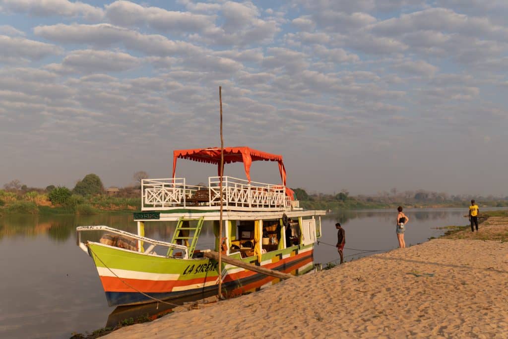 A small riverboat with a balcony on top moored on the edge of a river, next to a sandy island. Blue and white clouds streak across the sky.
