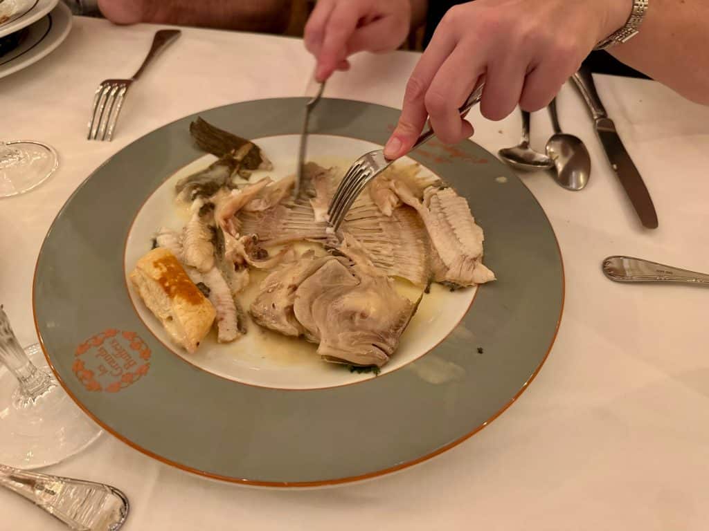 Two hands removing the bones from a flat fish dish with a fork and knife.