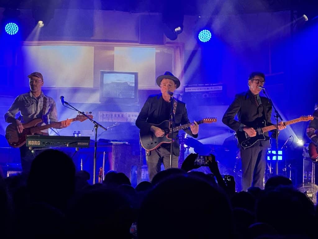 Belle and Sebastian the band playing on stage: three guitar and bass players singing at microphones.