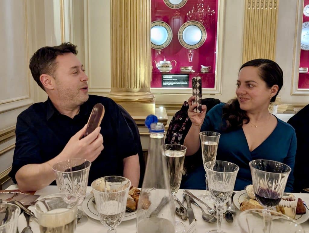 Charlie holds up a chocolate eclair and Kate takes a photo with her phone. They're surrounded by crystal goblets and silverware at Les Grands Buffets.