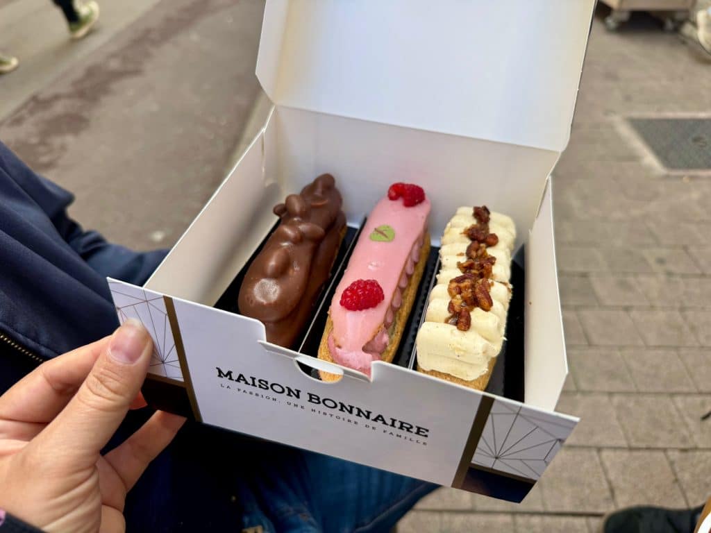 A box full of three eclairs: one chocolate with nuts and caramel, one raspberry, and one vanilla with pecans.