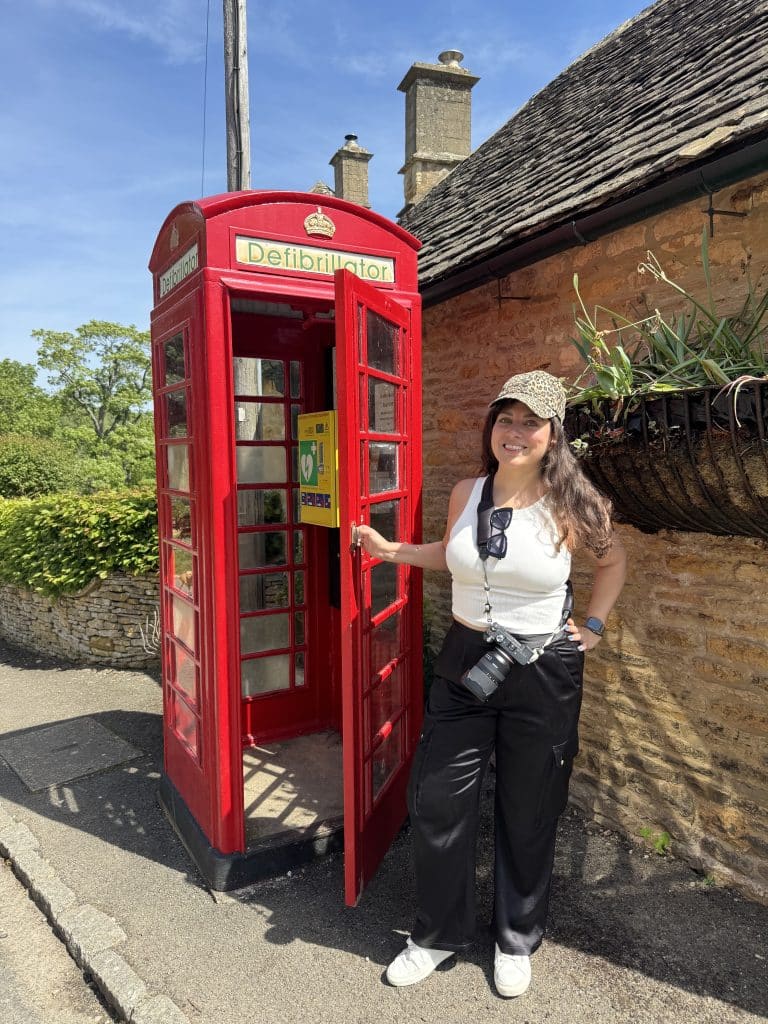 Kate berdiri di depan bilik telepon tradisional Inggris berwarna merah, yang sekarang berisi defibrilator, bukan telepon.