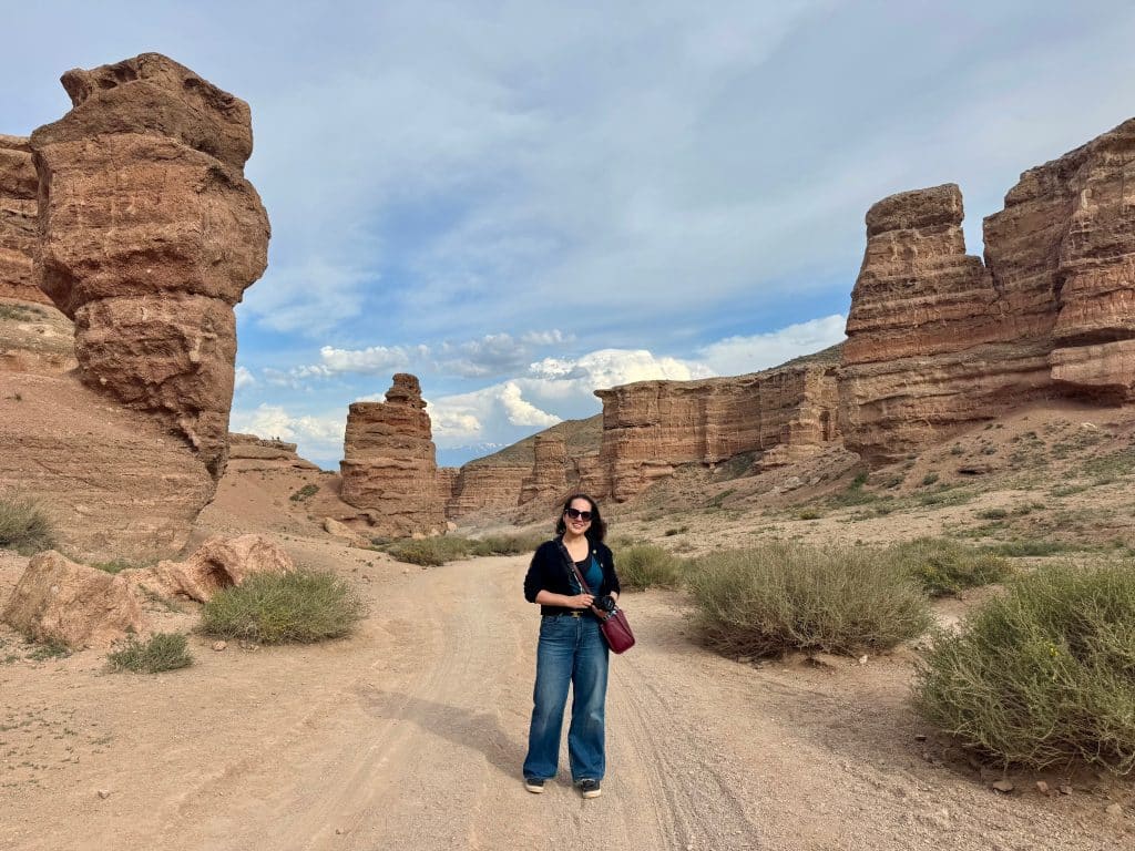 Kate standing on a dirt road with big sand-colored rock formations rising on either side of her.
