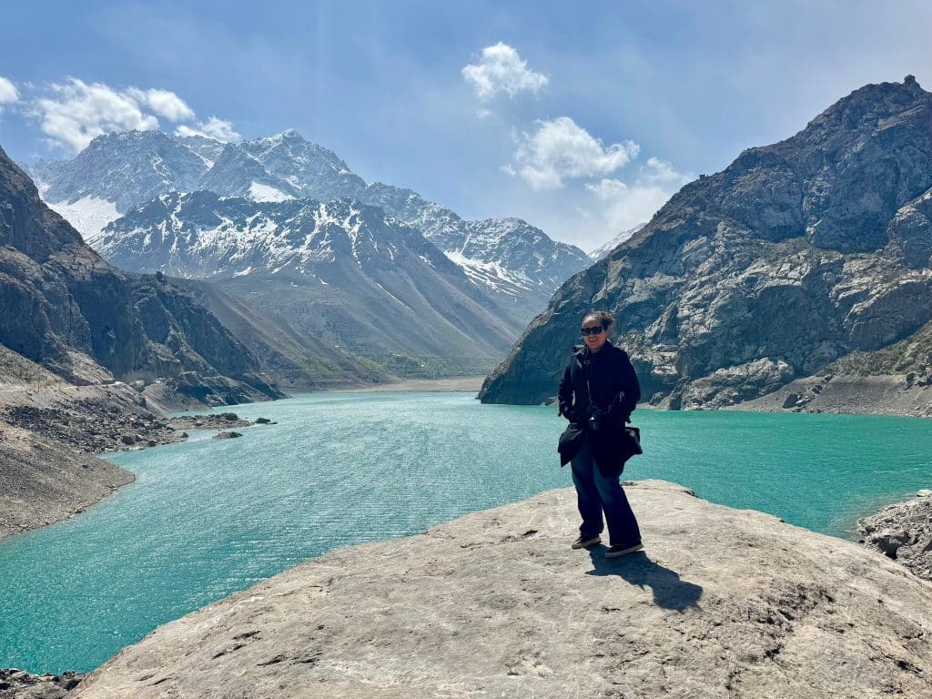 Kate in a black trench coat standing on a big, flat rock in front of a turquoise lake and snow-covered mountains.