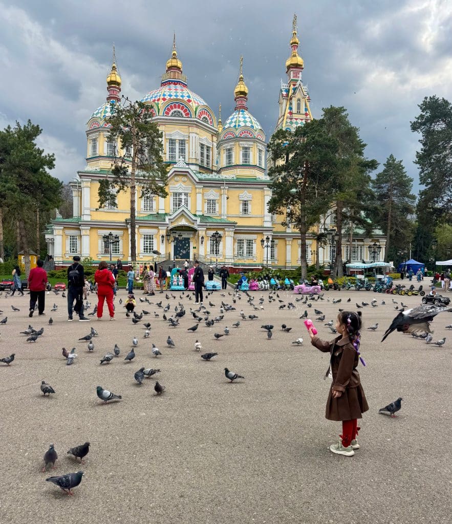 A little girl standing on a square in front of a church, pointing a bubble gun at pigeons.