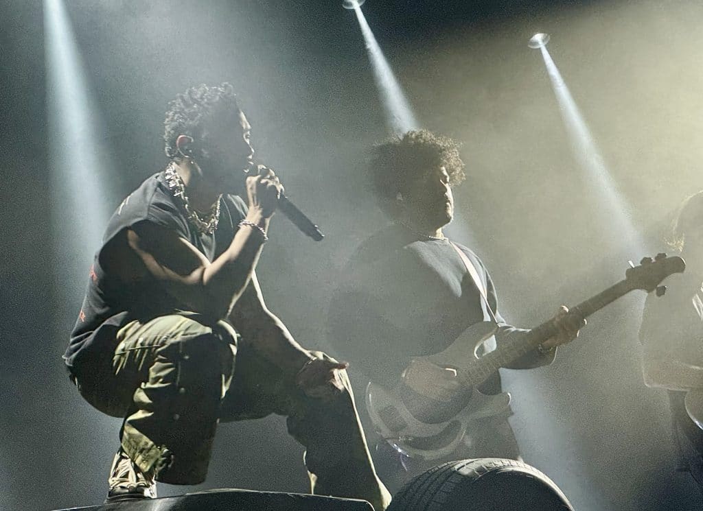Singer Miguel performing on stage, squatting while singing into a microphone, his bassist behind him.