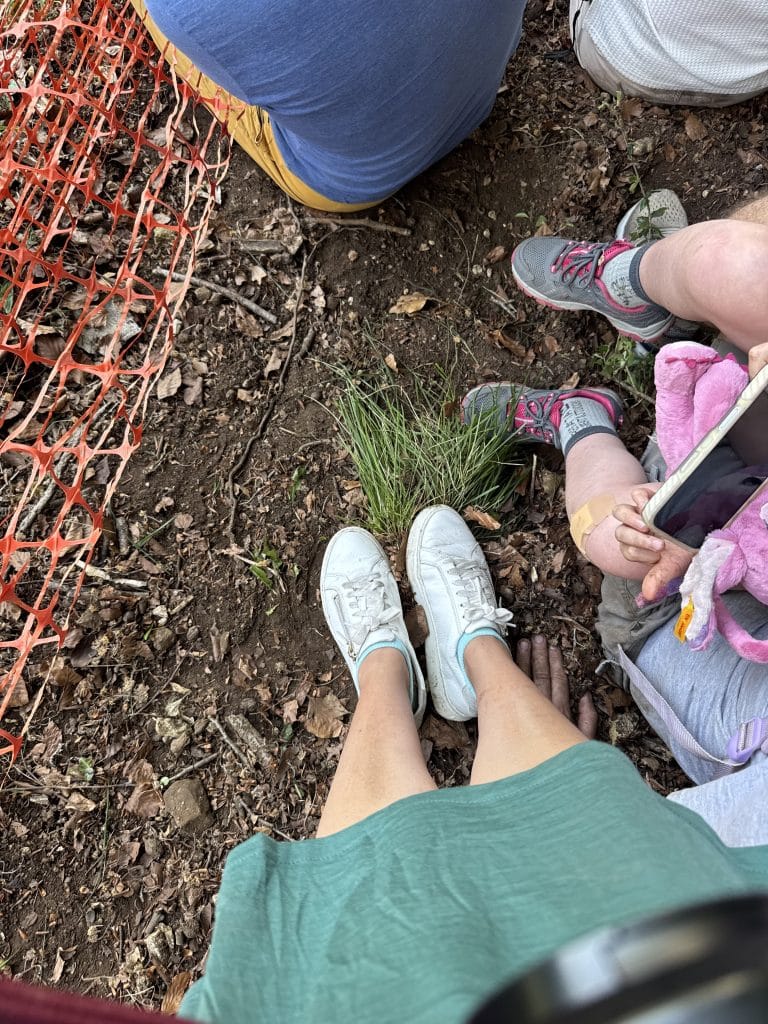 Kaki sepatu kets putih Kate bertengger di seberkas rumput kecil, dikelilingi tanah.