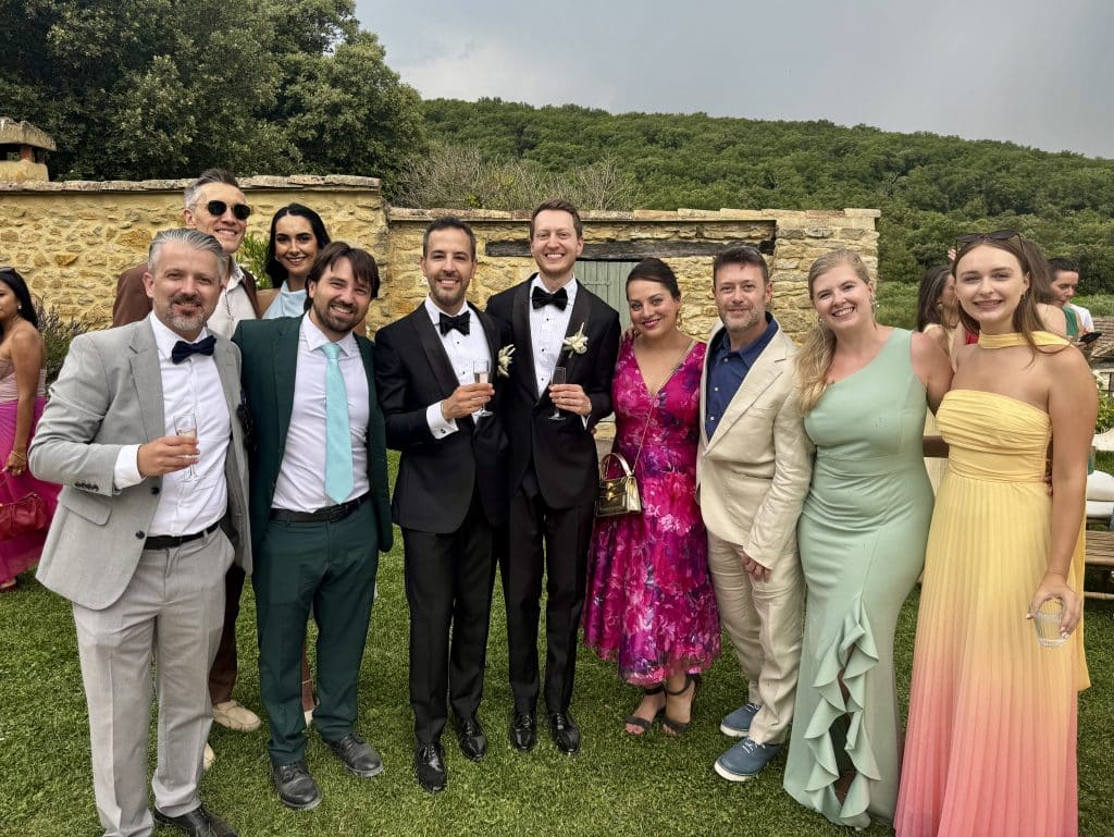 Kate, Charlie, and several friends in gowns and suits with the two grooms in the center in black tuxedos.