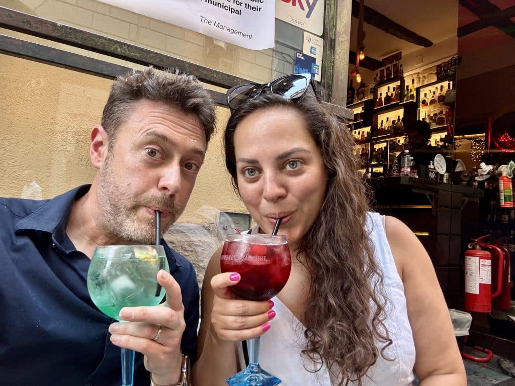 Kate and Charlie taking a selfie while sipping from spritzes in Naples. His is turquoise, hers is dark red.