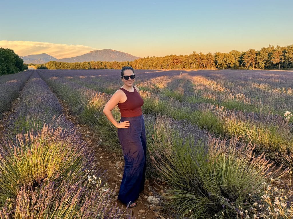 Kate standing in a field of lavender at sunset, wearing a burgundy tank top and navy wide legged linen pants.