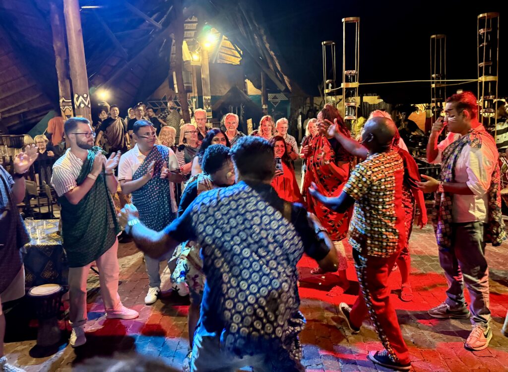 A group of Africans and tourists dancing together, wearing patterned pieces of cloth tied on top of their clothes.