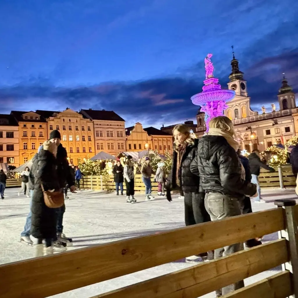 People skating on a rink on a square in front of bright yellow buildings beneath a darkening blue sky.