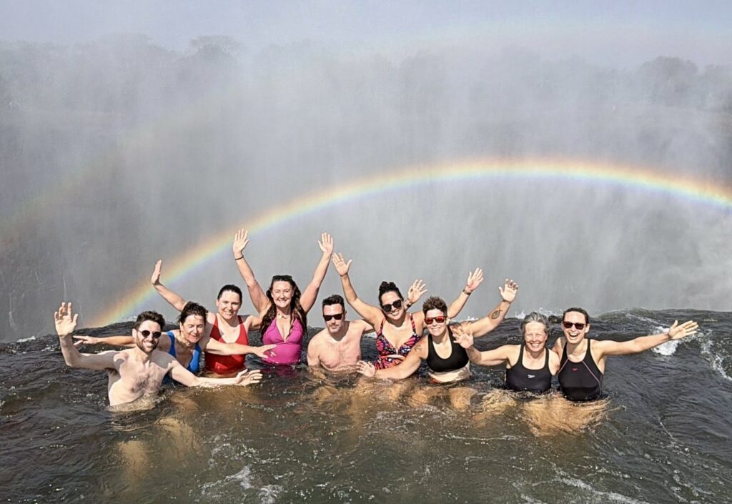 Kate, Charlie, and seven of their friends posing on the edge of the Devil's Pool waterfall with a double rainbow in the mist behind them.