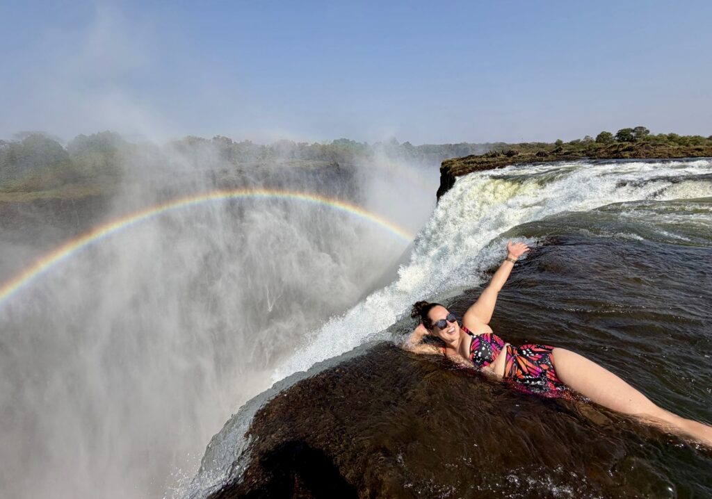 Kate lying on the edge of Victoria Falls, like she's about to go over, but she strikes a glamorous pose on her side. There is a rainbow in the mist behind her.