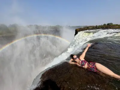 Kate lying on the edge of Victoria Falls, like she's about to go over, but she strikes a glamorous pose on her side. There is a rainbow in the mist behind her.