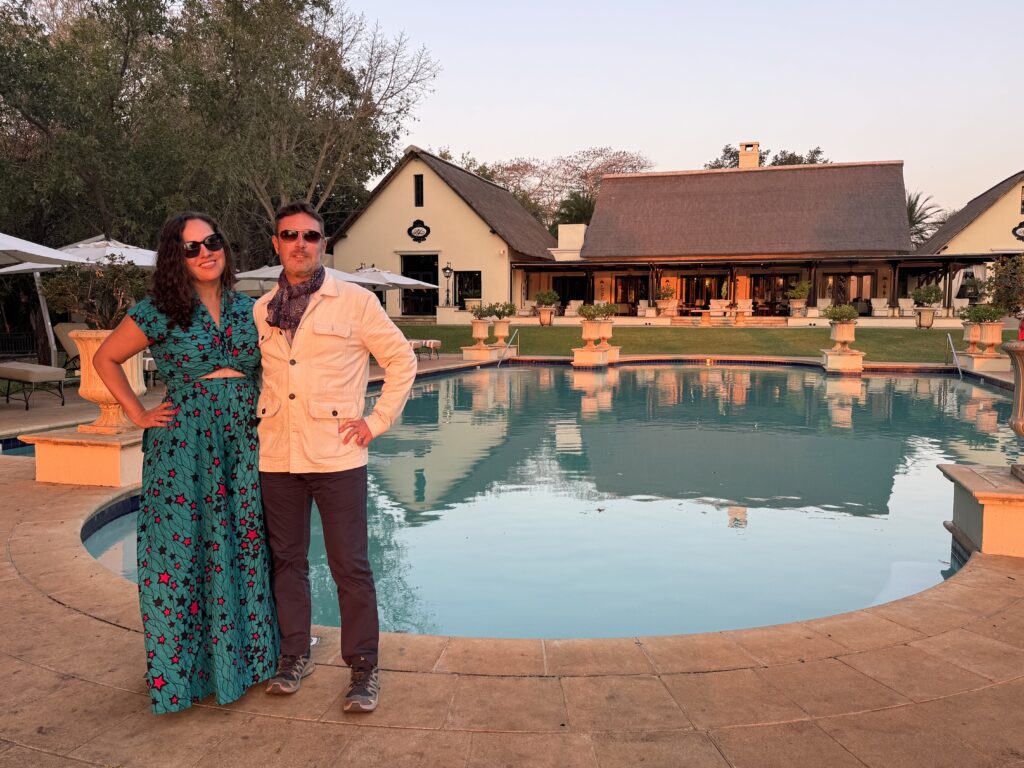 Kate and Charlie posing in front of a pool at a luxury hotel. Kate wears an African outfit: a two-piece top and wide-leg trousers in a turquoise pattern with hot pink stars. Charlie wears a khaki jacket, patterned navy scarf, and navy trousers.