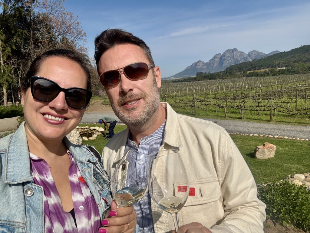 Kate and Charlie toasting tasting glasses of white wine in Franschhoek, South Africa, with blue mountains and green vineyards behind them.