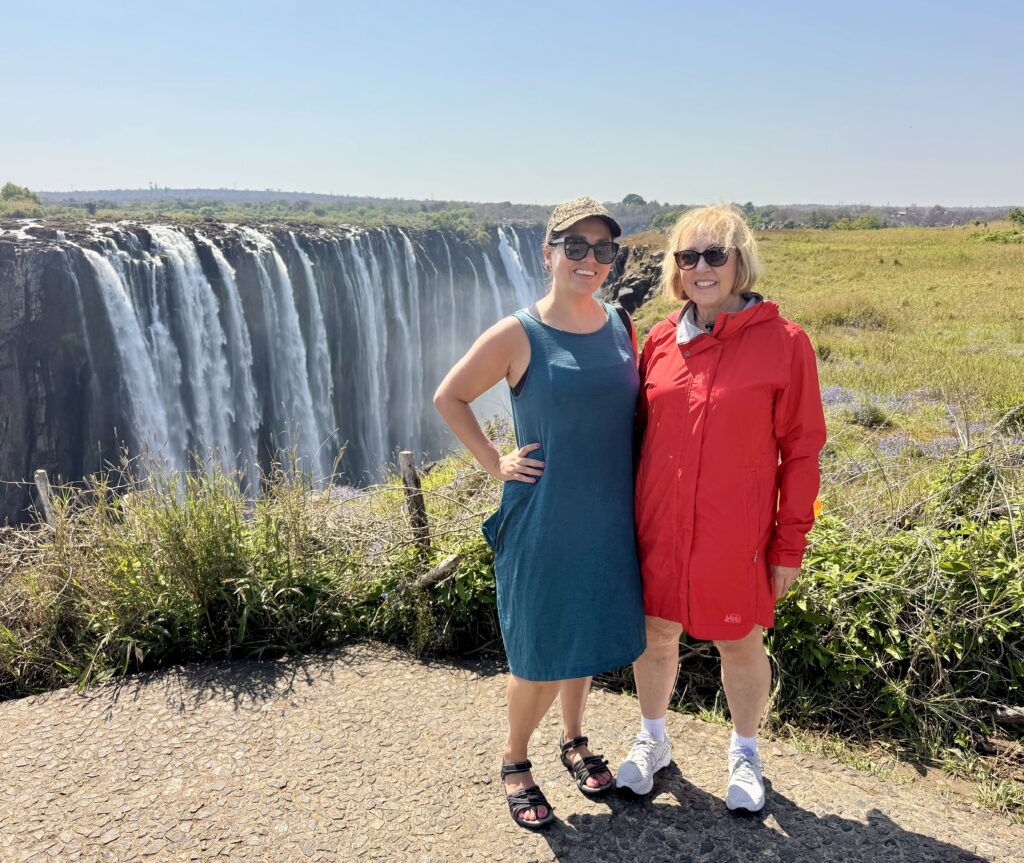 Kate and her mom standing arm and arm with Victoria Fall's multi-tiered waterfalls behind them.